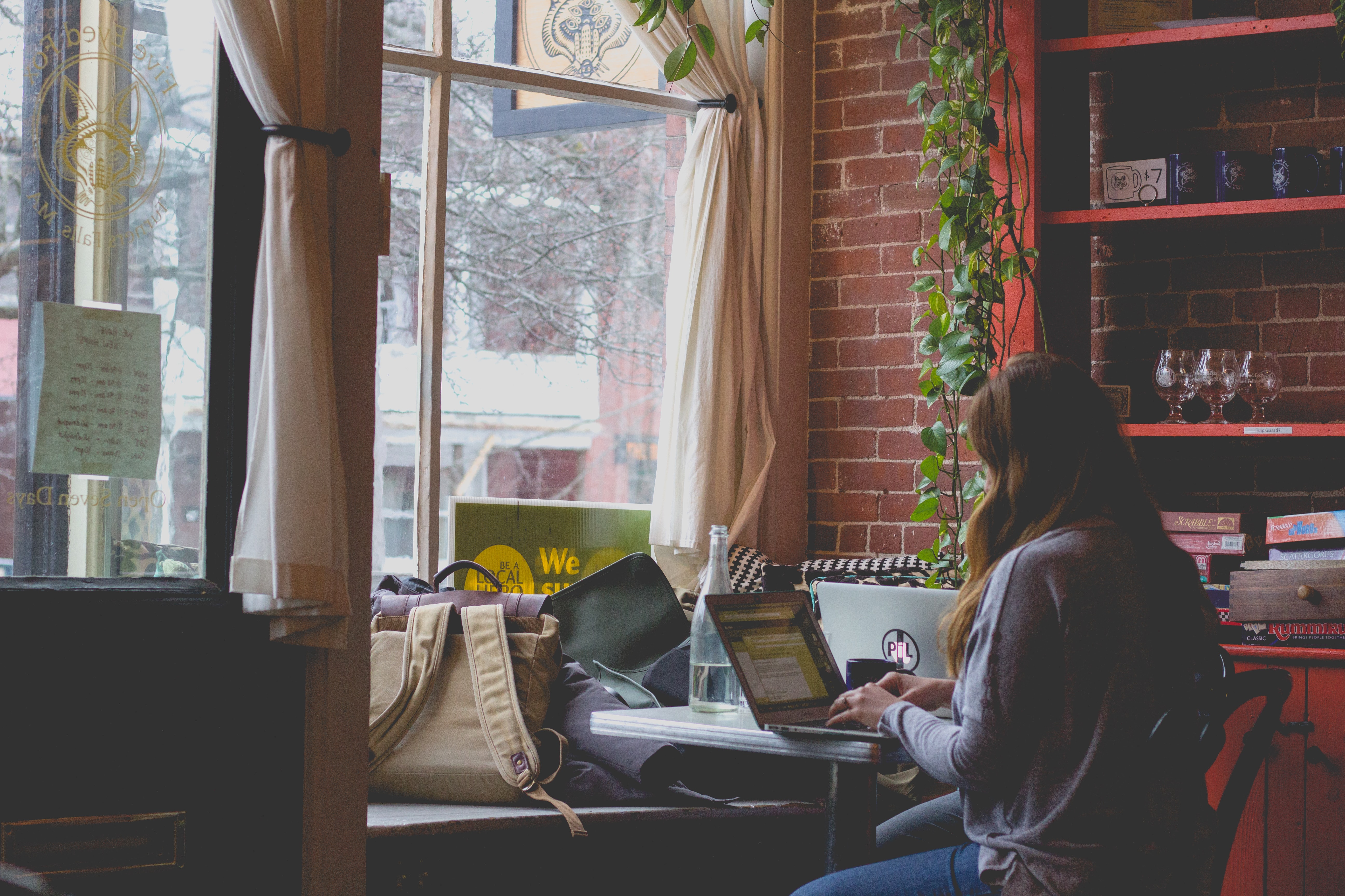 The back of a young woman sitting at a desk on her laptop in front of a brick wall